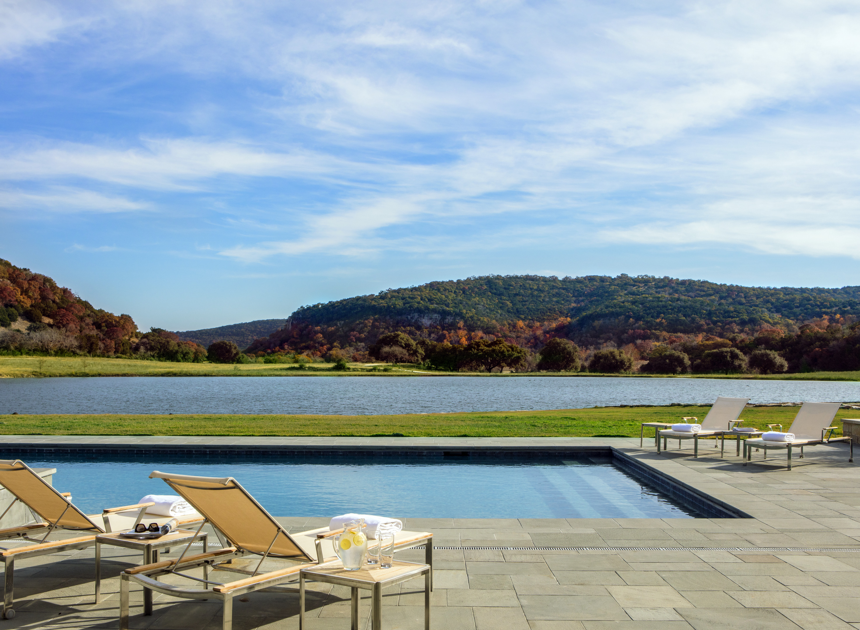 The outdoor swimming pool at Skull Creek Ranch features inviting lounge chairs and towels, all set against a stunning backdrop of hills and a shimmering body of water beneath a partly cloudy sky.