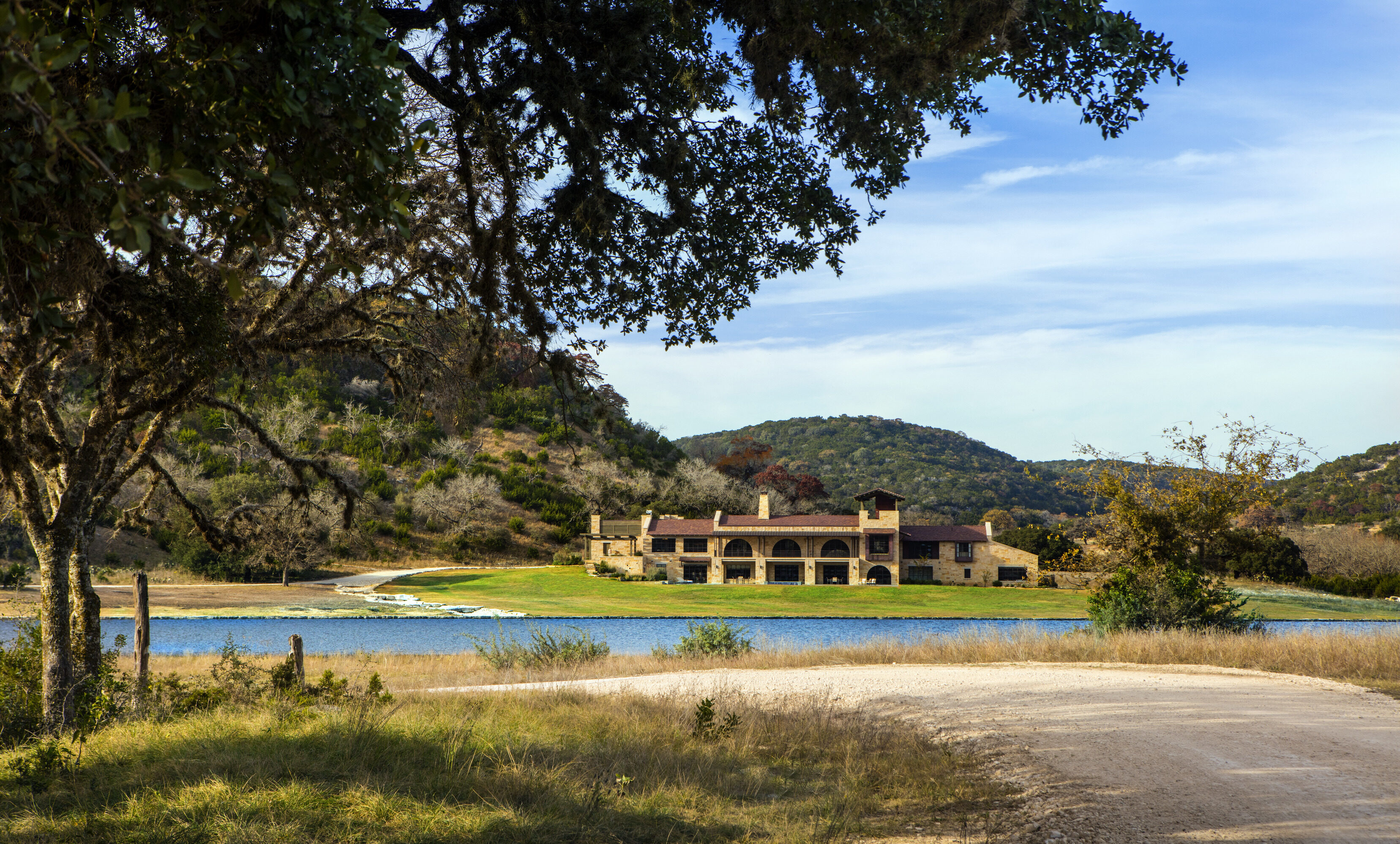 A countryside house by Skull Creek Ranch sits by a river, surrounded by trees and hills under a blue sky. A dirt road gently winds its way toward the building, inviting exploration.