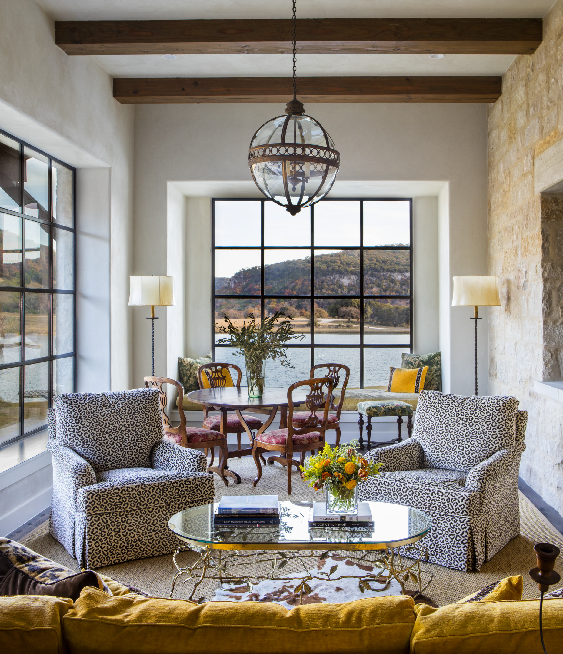 The living room at Skull Creek Ranch features patterned armchairs, a glass coffee table, and a cheerful yellow sofa. Large windows offer picturesque views of the lake and hills, while a round dining table and chairs sit gracefully in front of the window.