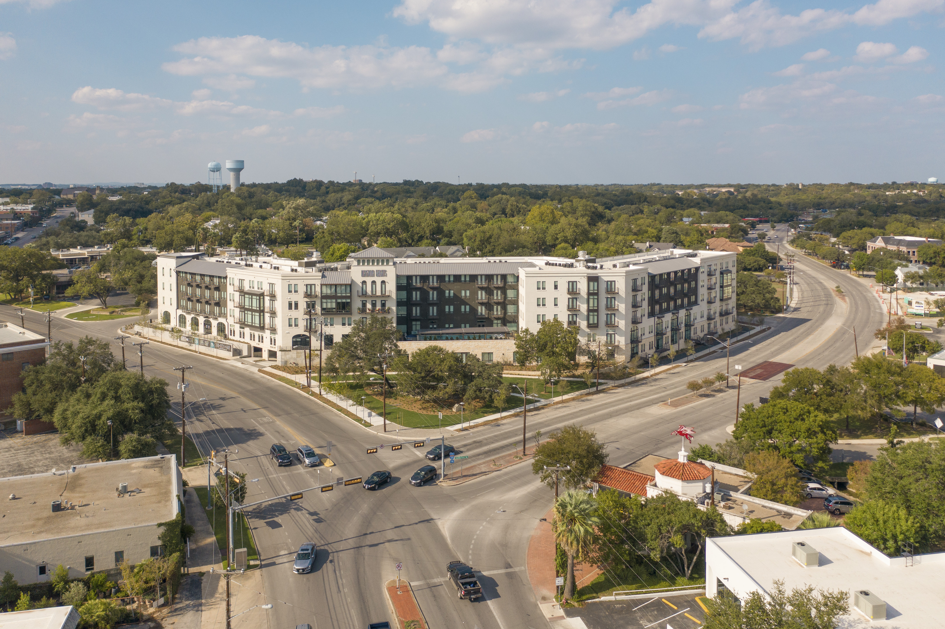 Aerial view of the bustling intersection at Magnolia Heights, showcasing cars weaving around a large, multi-story mixed-use development surrounded by lush trees and smaller structures on a sunny day.