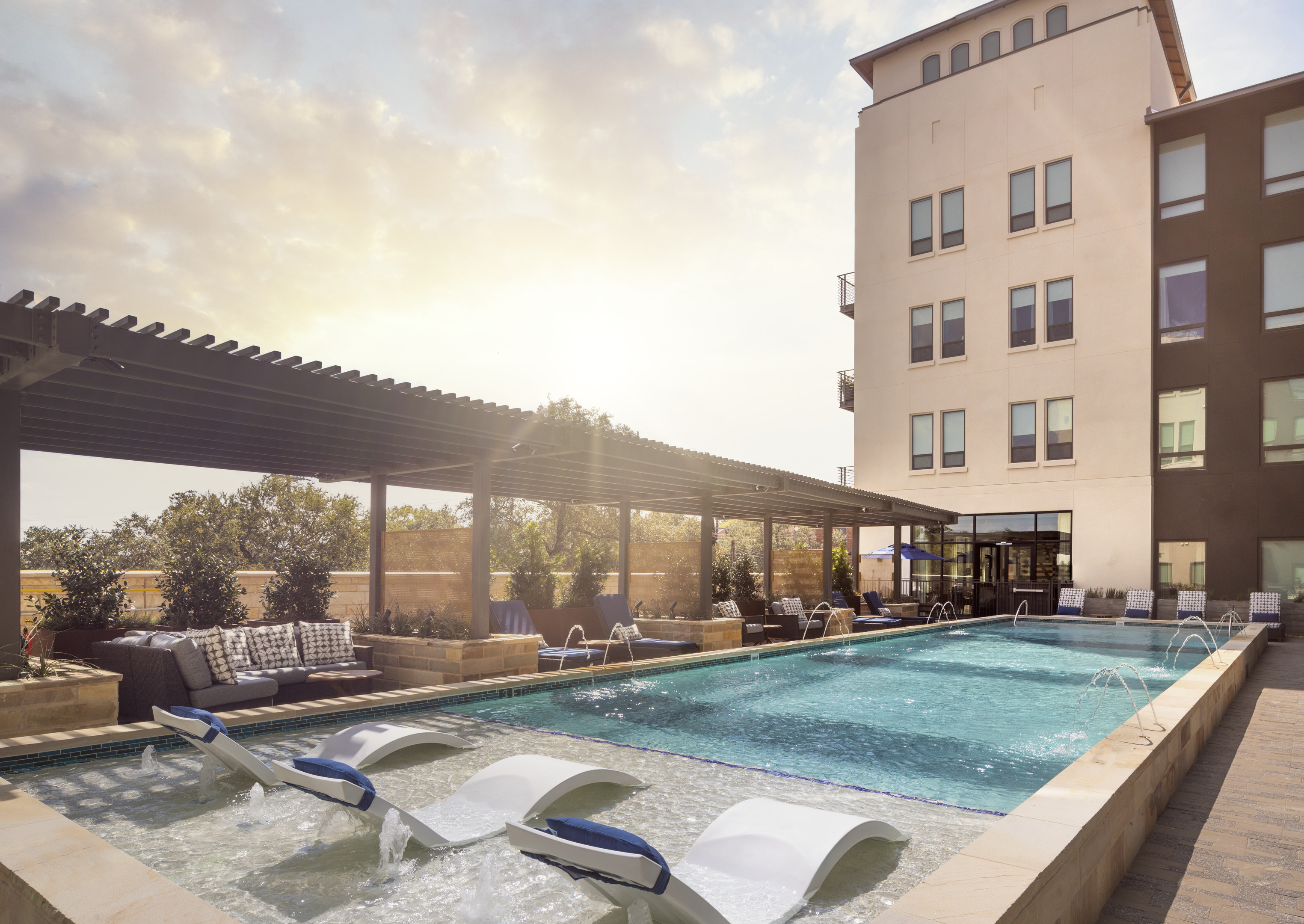 An outdoor swimming pool with lounge chairs and a shaded seating area sits gracefully beside Magnolia Heights, a stunning example of modern mixed-use development, under a partly cloudy sky at sunset.