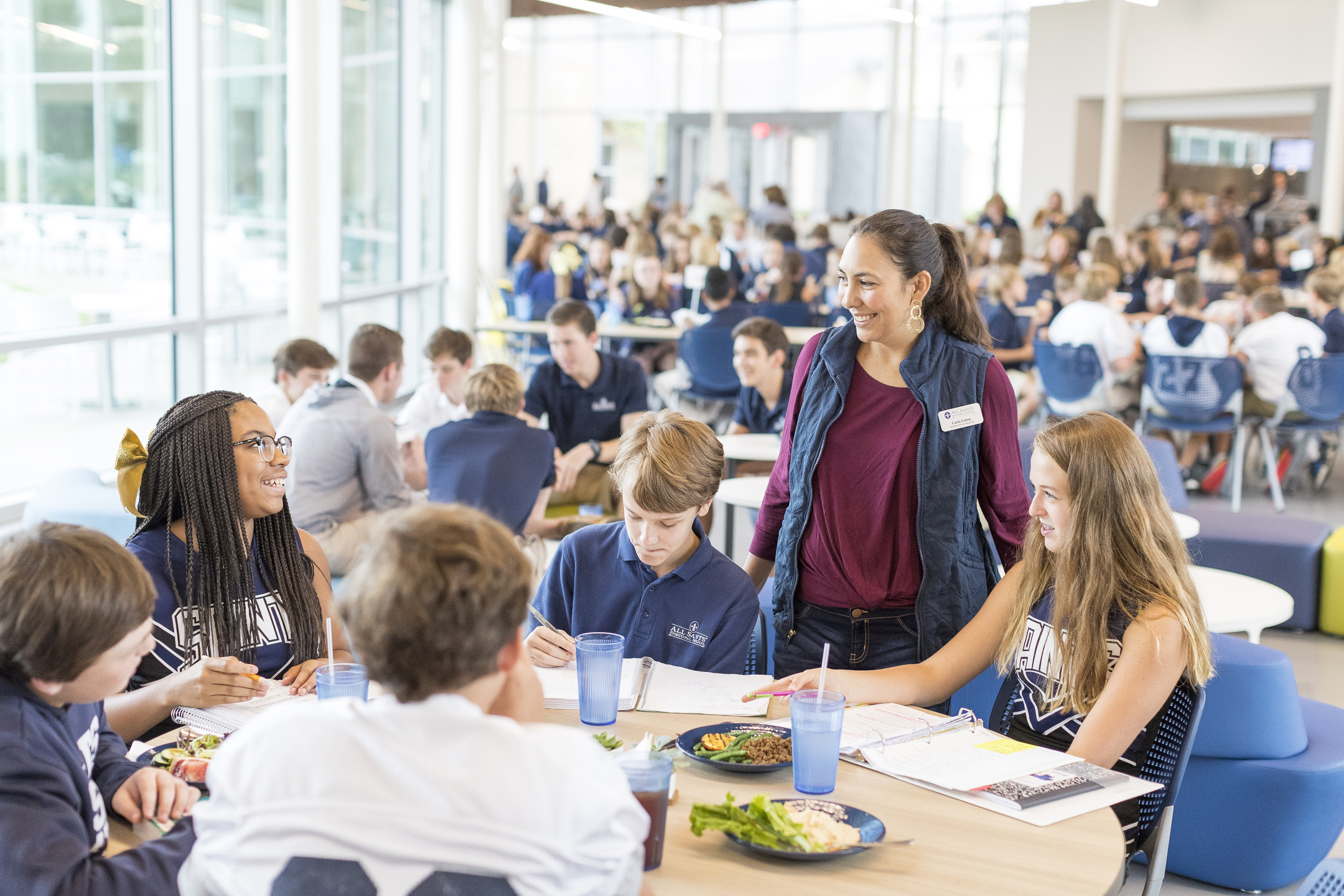 In a bustling cafeteria, a group of students sits around the table, chatting and eating as an adult stands nearby, engaging in their lively Auto Draft of conversation.