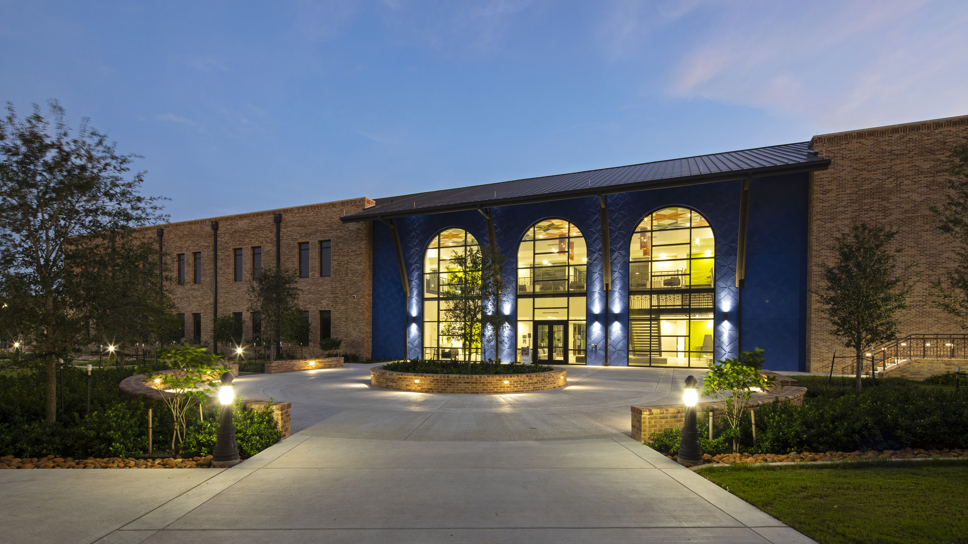 A modern brick building with large arched windows, illuminated at dusk, stands elegantly. A circular driveway and a meticulously landscaped area in front add to its charm, seemingly as perfect as an auto draft of architectural brilliance.