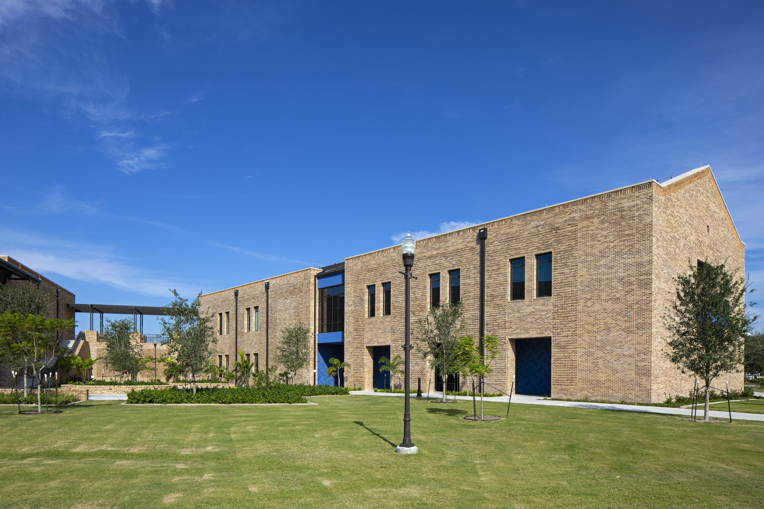 A modern brick building with large windows under a clear blue sky stands as if crafted by an auto draft, surrounded by a well-maintained lawn with a lone street lamp and young trees.