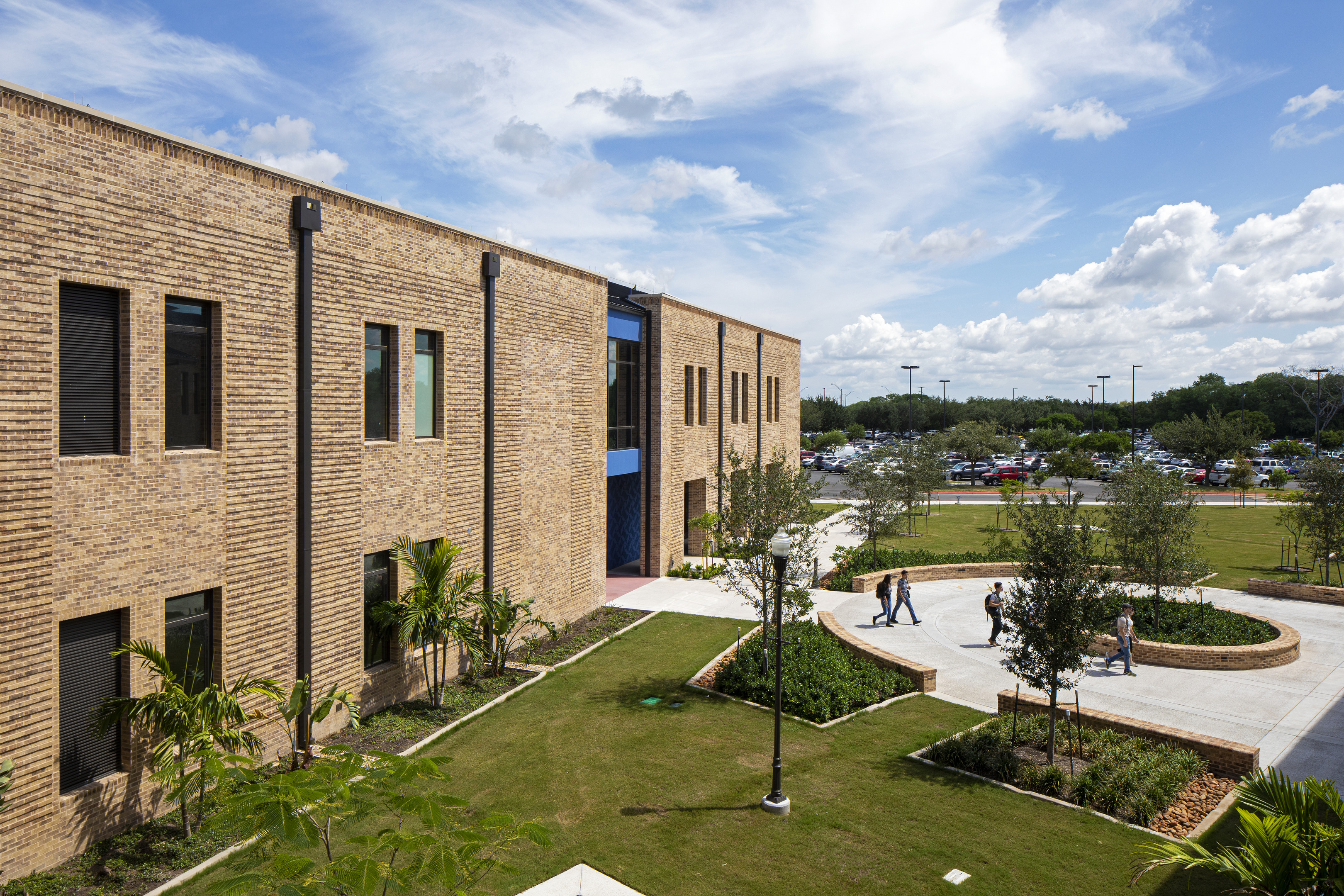 A modern brick building with a courtyard featuring grass, trees, and walkways under a partly cloudy sky. People stroll near the building while cars are smoothly parked in the lot, showcasing an auto draft of daily life in motion.