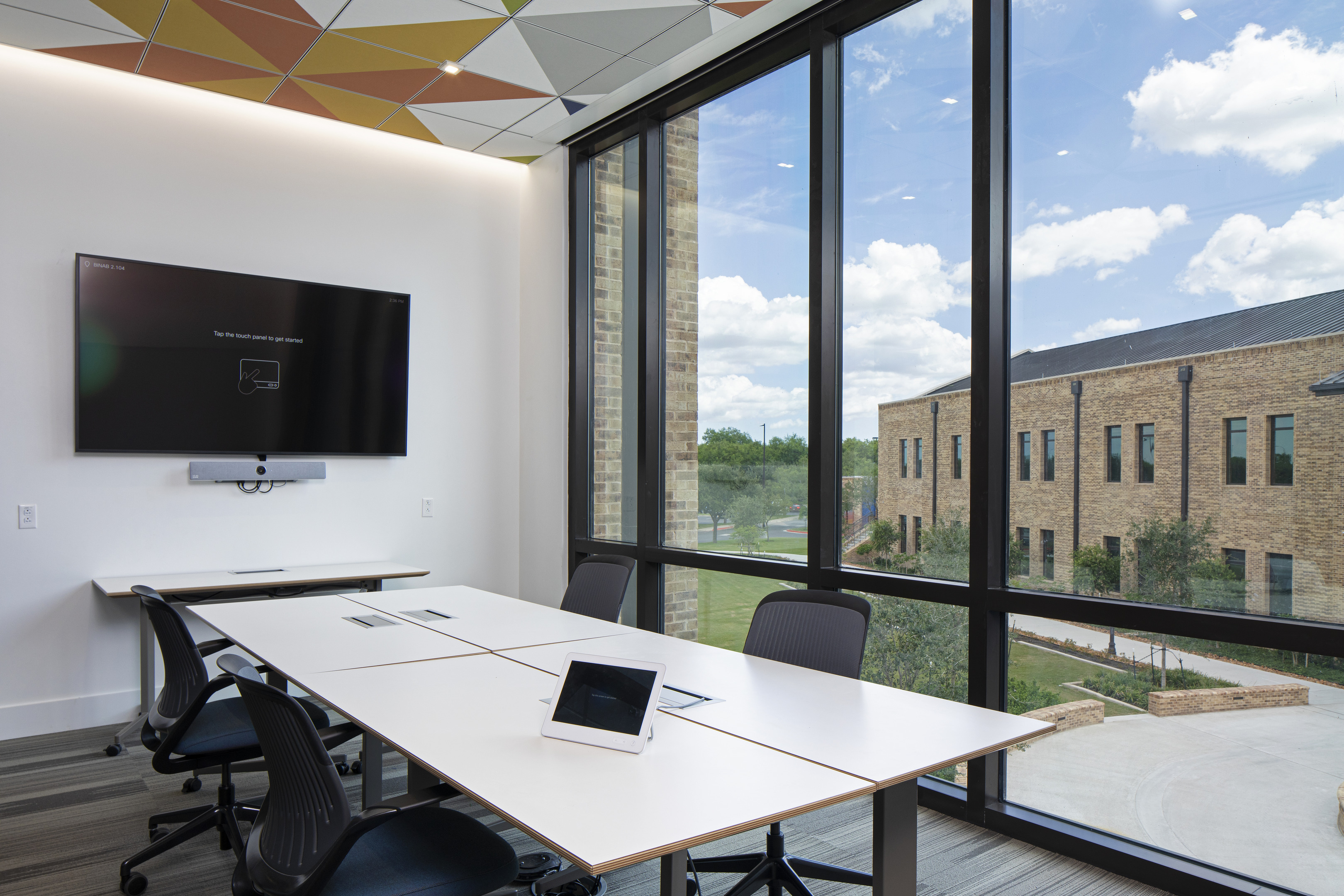 A modern conference room equipped with a long table, chairs, and a wall-mounted screen for seamless presentations creates an ideal space for collaboration. A tablet enables auto draft note-taking, while large windows offer views of a brick building against a cloudy sky.