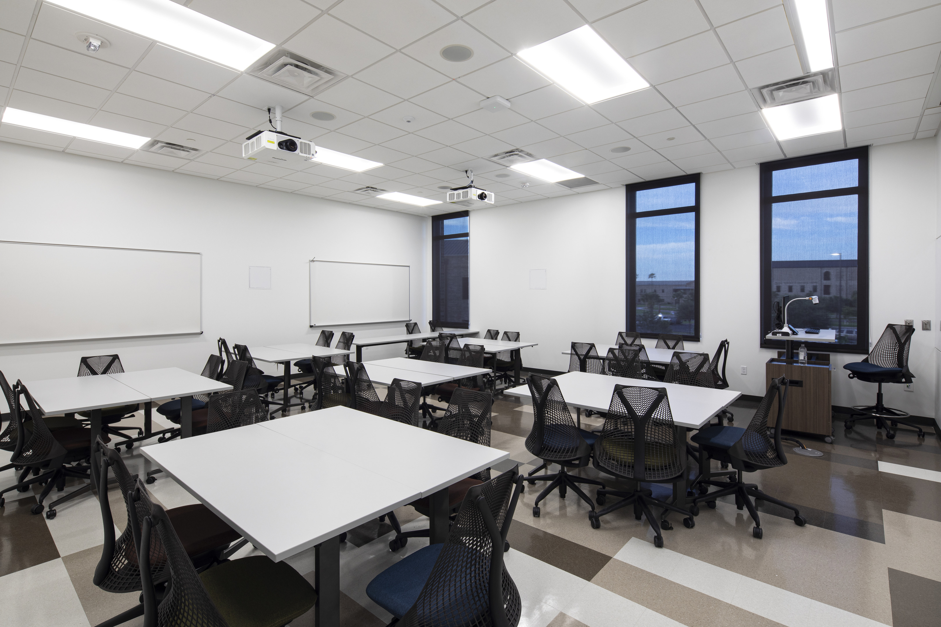 A modern classroom with several tables and chairs, three projectors, and large windows offers an ideal space for service-oriented learning. Whiteboards are mounted on the walls, while the floor showcases a pattern of light and dark tiles.