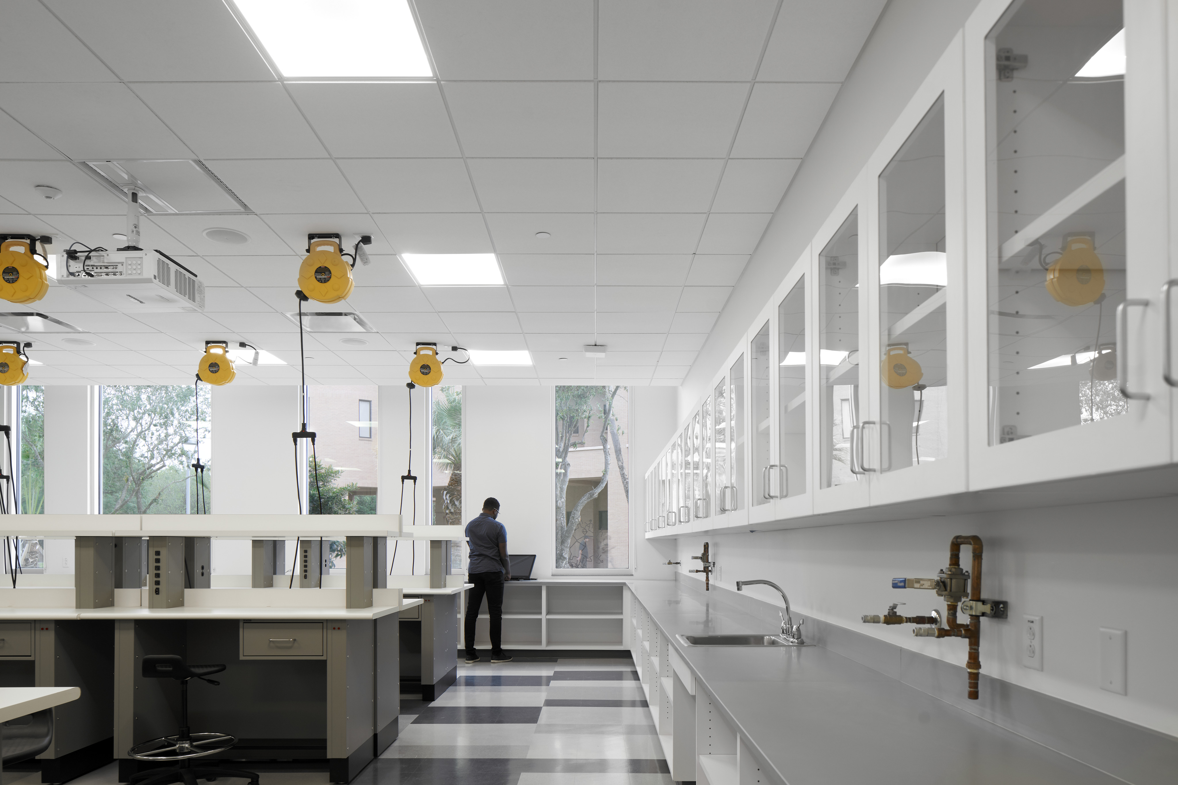 A person stands at a workstation in a modern laboratory with auto draft capabilities, surrounded by white cabinets, overhead equipment holders, and large windows.