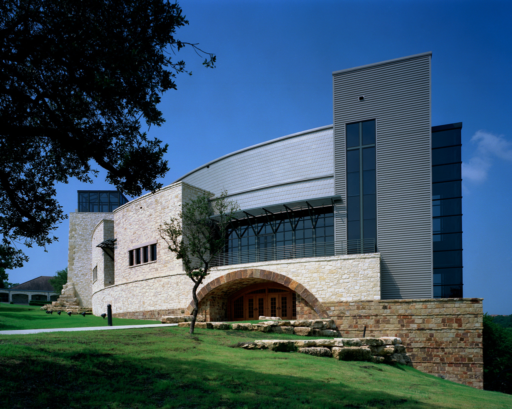 The Riverbend Church is a modern building with a stone and metal facade, featuring large glass windows and an arched entrance. Set on a grassy hillside under a clear blue sky, it stands majestically as a harmonious blend of nature and architectural ingenuity.