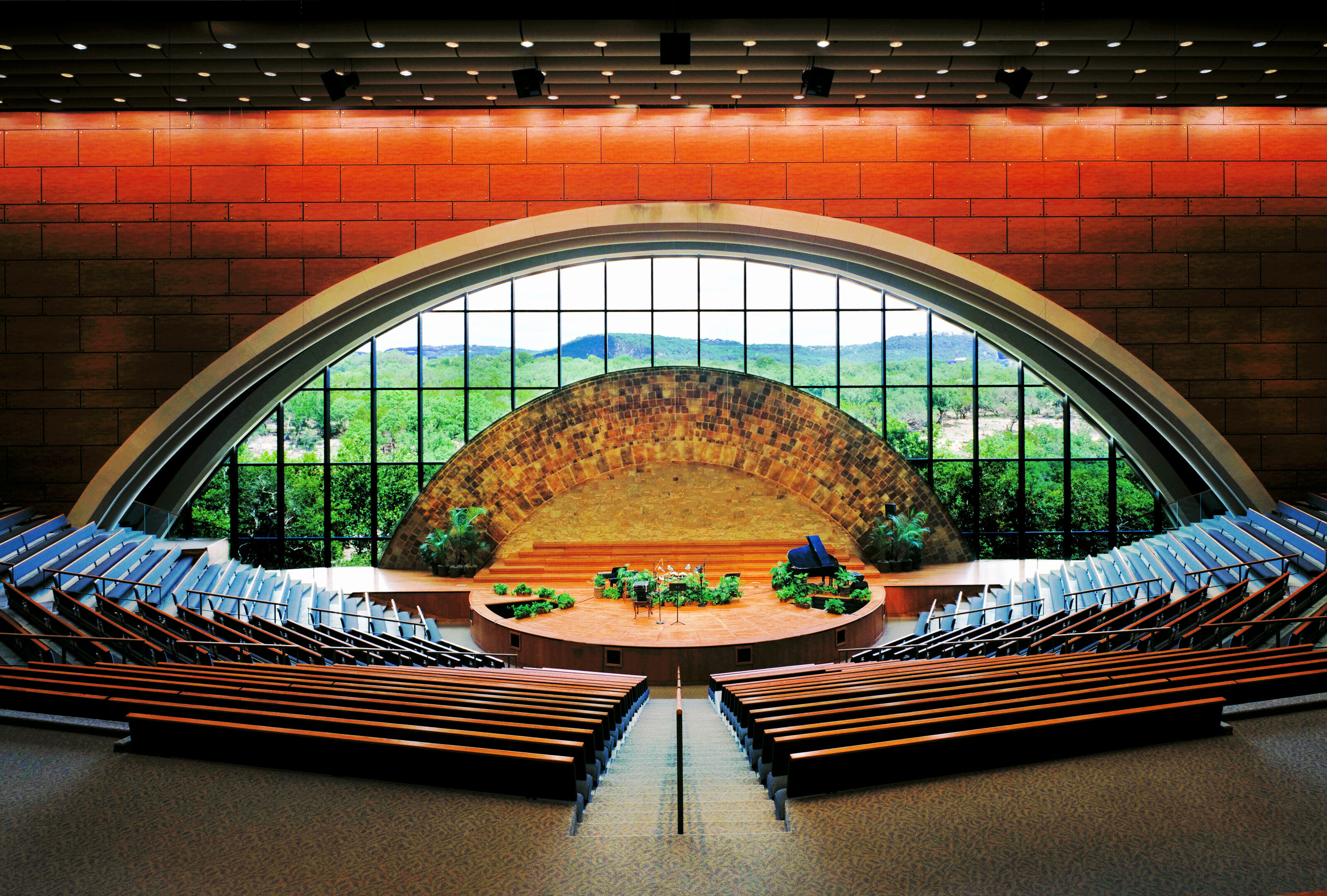 The auditorium at Riverbend Church features wooden seating, all facing a stage backed by a large, semi-circular window that frames a breathtaking view of greenery and distant mountains.