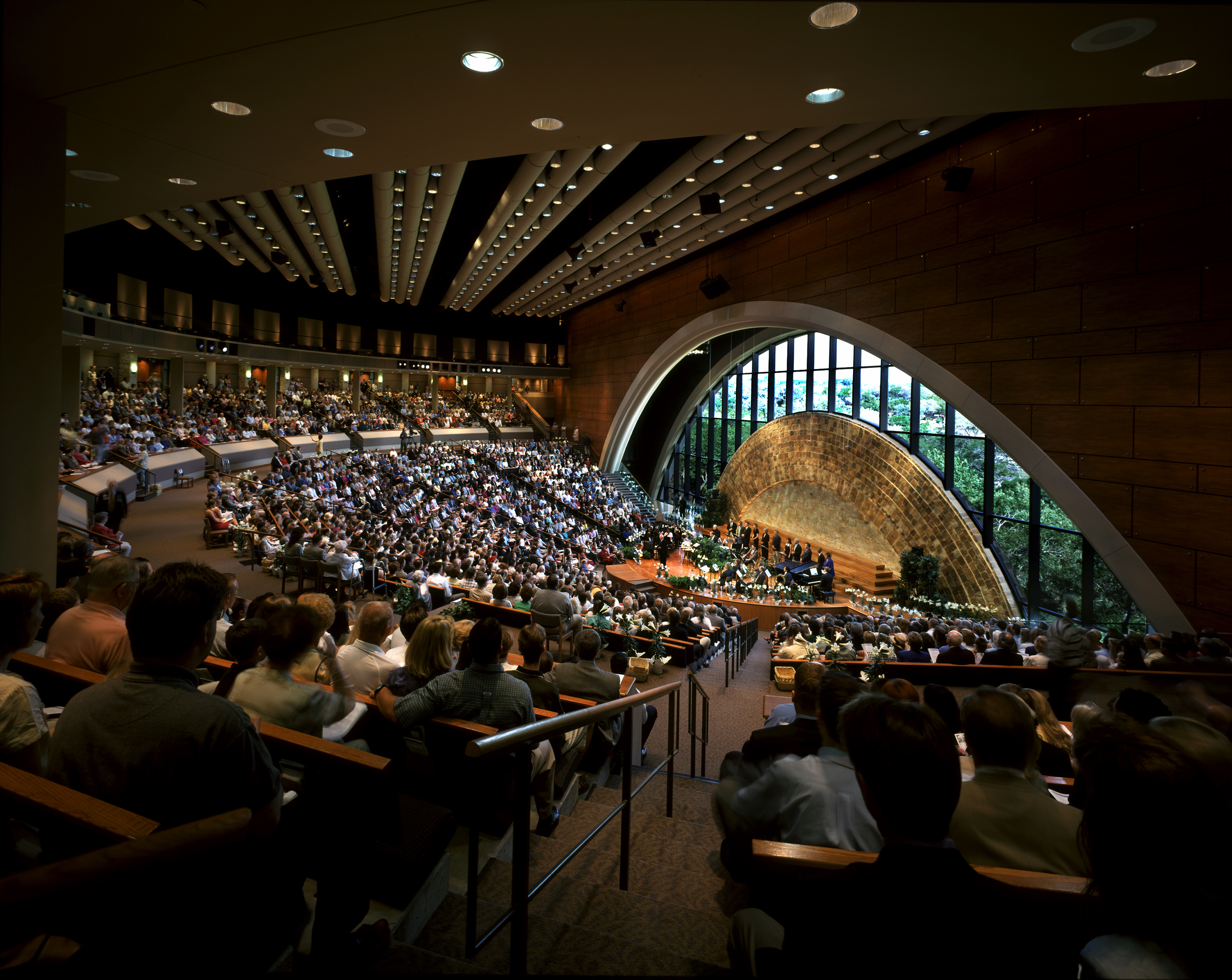 A crowded auditorium at Riverbend Church, featuring a large arched window and wooden ceiling, hosts an event with people seated and speakers on stage.