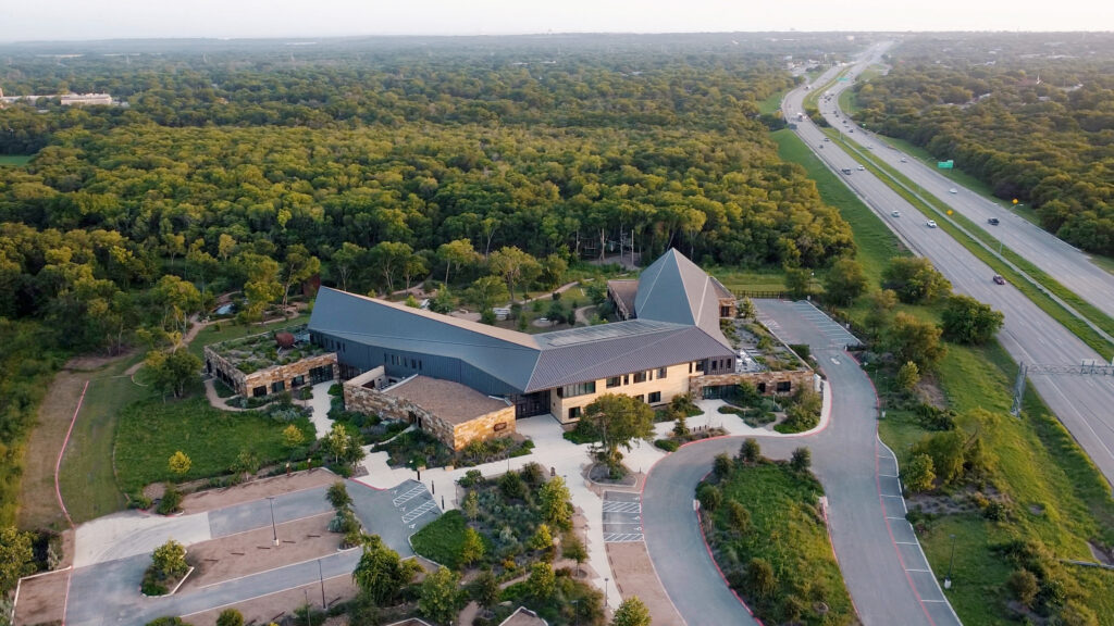 Aerial view of the Salado Creek campus, a modern, angular building embodying trauma-informed design principles. Nestled in a forest with a nearby highway, the ChildSafe facility boasts visible parking spaces and pathways for easy access.