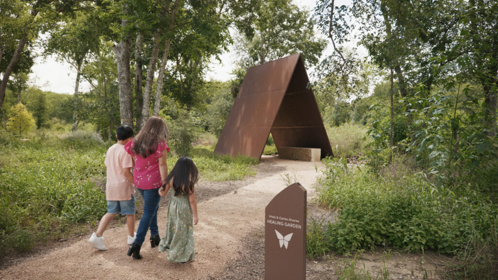 Three children walk towards a triangular rust-colored sculpture in a lush garden at the ChildSafe Salado Creek campus, with a sign labeled "Healing Garden" nearby. The space is thoughtfully crafted following trauma-informed design principles to offer tranquility and comfort.