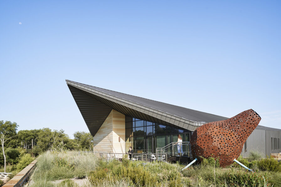 A modern building with a sharp, angular roof and glass walls stands on the serene Salado Creek campus, surrounded by greenery. A rust-colored geometric sculpture rests in the foreground under a clear blue sky, exemplifying trauma-informed design.