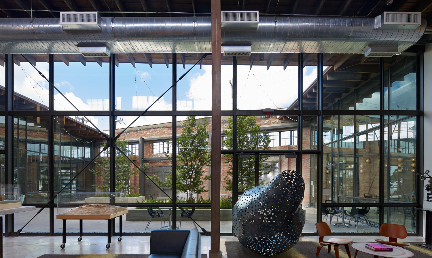 Modern industrial-style office space with large glass windows, exposed ductwork, indoor plants for sustainability, a sculptural art piece, and views of an outdoor courtyard.